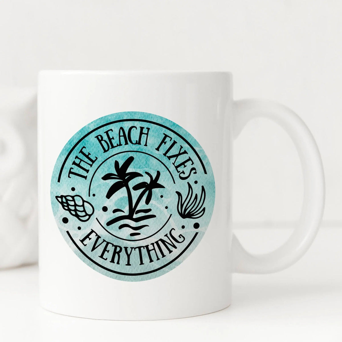 White ceramic coffee mug with teal beach logo and curved text “The Beach Fixes Everything”