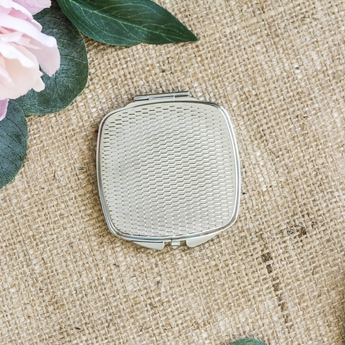 Silver square compact mirror with textured white back, closed on woven fabric.