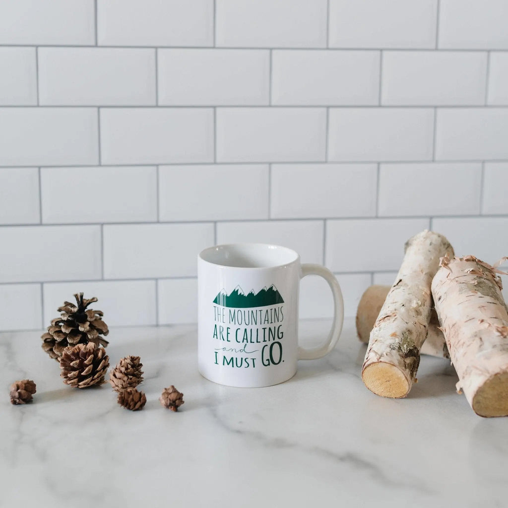 White ceramic coffee mug with green mountain quote and pinecone decor nearby