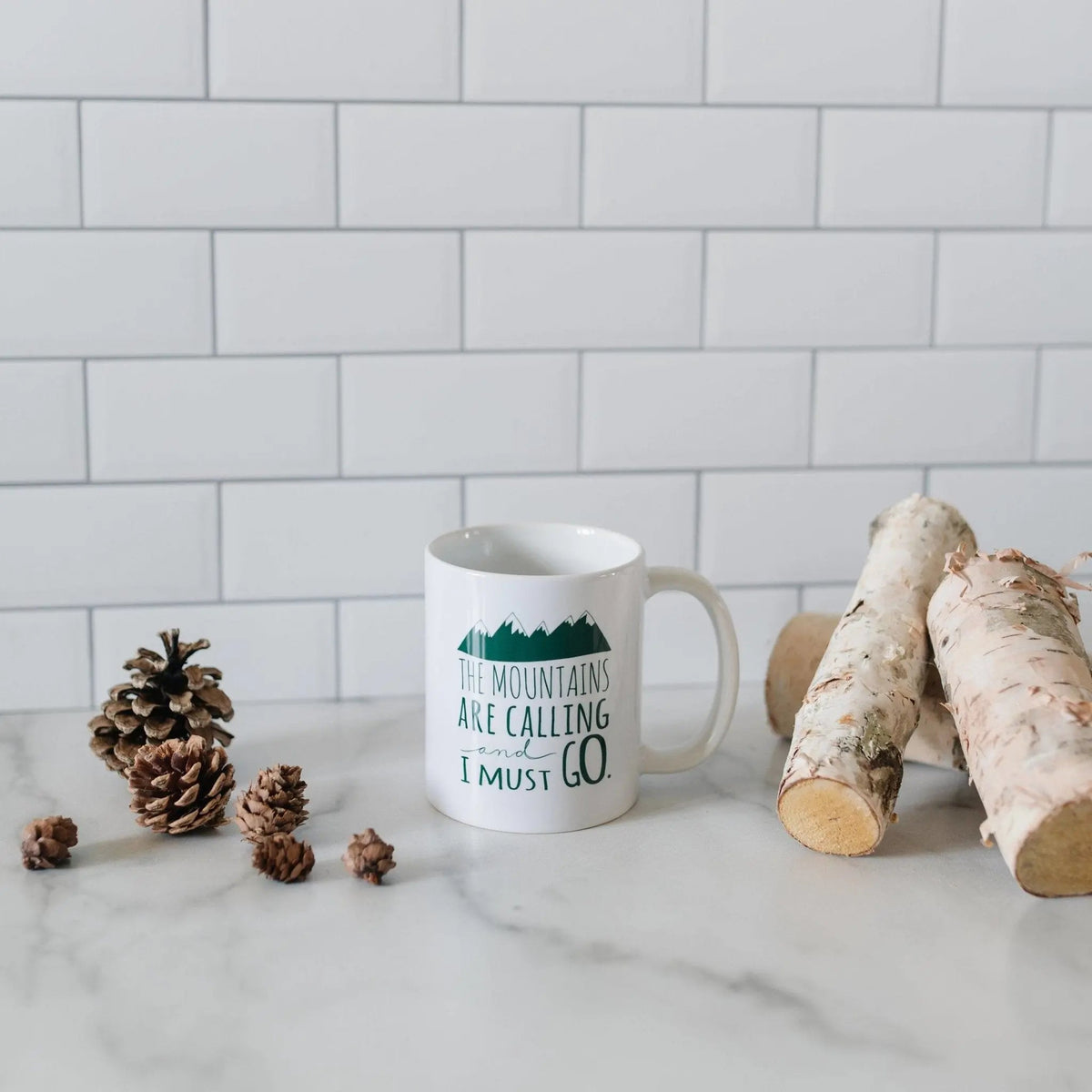 White ceramic coffee mug with green mountain quote and pinecone decor nearby