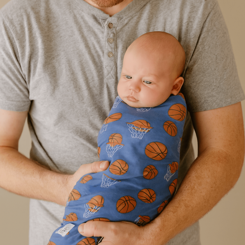 Blue basketball hoop swaddle blanket with hoop and ball pattern, worn on baby.